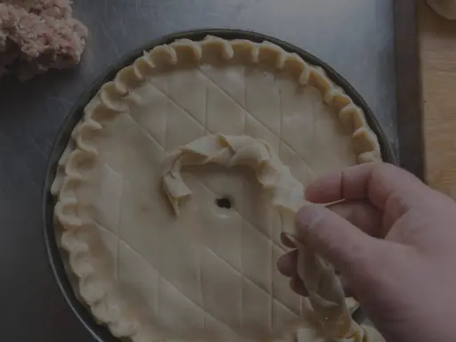 Main d'un pâtissier décorant une tarte avec une pâte brisée tressée. Un tas de farce est visible à gauche.