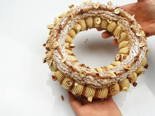 Un Paris-Brest craquelin, orné de noisettes et de caramel, est tenu par des mains.