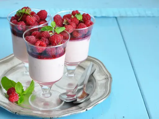 Trois panna cotta onctueuses, garnies de framboises fraîches et d'un coulis de framboise, sur un plateau argenté.