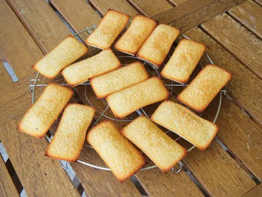 Douze biscuits financiers dorés, disposés sur une grille de refroidissement, sur une table en bois.