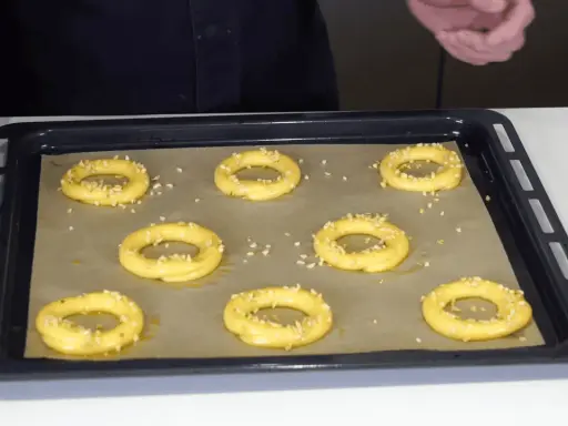 Huit ronds de pâte à choux, prêts à devenir un délicieux Paris-Brest, sont saupoudrés d'amandes effilées sur une plaque de cuisson.