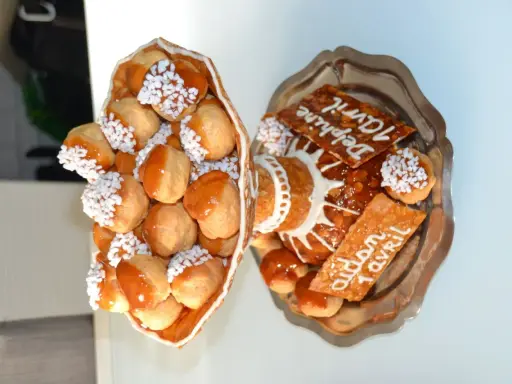 Une pièce montée de choux garnis de crème, nappés de caramel et décorés de perles blanches. Sur un plateau, d'autres choux et des tuiles aux amandes portent des inscriptions.