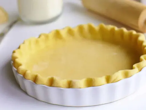 Pâte brisée prête pour la cuisson dans un plat à tarte blanc, avec un rouleau à pâtisserie en arrière-plan.