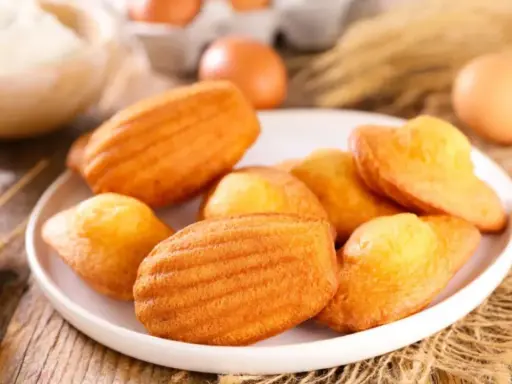 Des madeleines dorées, parfaites pour une recette gâteau madeleine meilleur pâtissier. Des œufs et de la farine en arrière-plan.