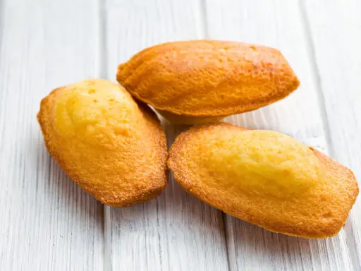 Trois madeleines dorées, tout juste sorties du four, sur une planche en bois. La cuisson madeleine est réussie, leur texture est parfaite.