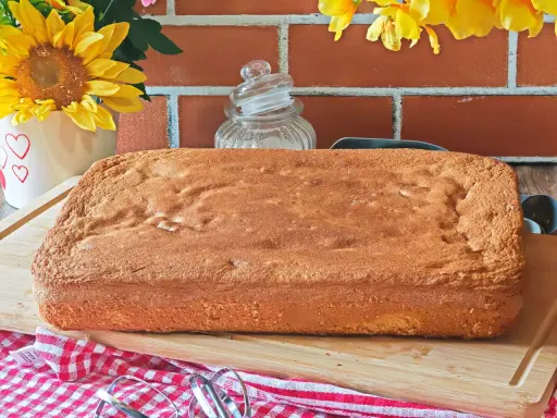 Une génoise rectangulaire dorée, parfaite pour 15 personnes, attend d'être garnie. Des fleurs jaunes et un fond de briques rouges créent une ambiance chaleureuse.