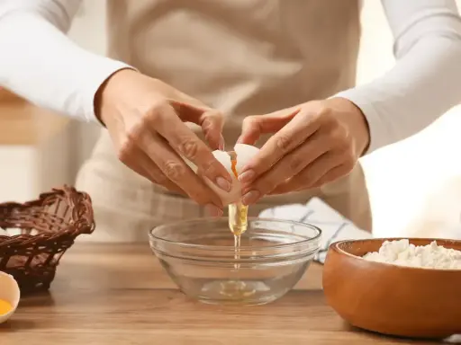 Préparation d'une recette pâte brisée facile : une main casse un œuf dans un bol, à côté d'un panier d'œufs et d'un bol de farine.
