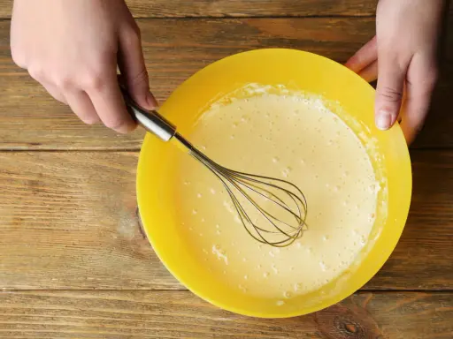 Mains préparant la pâte à crêpes dans un bol jaune. La pâte est prête pour son temps de repos.