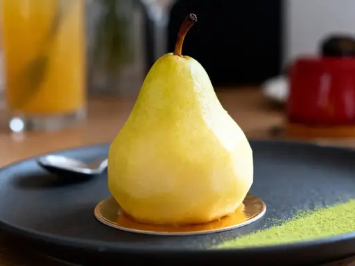 Un entremet poire, glacé et brillant, repose sur une assiette noire avec une touche de poudre verte.