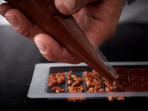 Main d'un artisan versant du chocolat fondu dans un moule pour créer une tablette de chocolat maison garnie de noisettes caramélisées.
