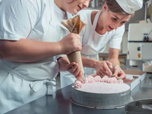 Deux pâtissières décorent un gâteau rose lors d'un cours de pâtisserie en ligne. L'une utilise une poche à douille, l'autre des fruits.