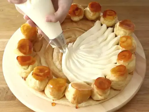Une main garnit une couronne de choux avec de la crème chiboust, créant un dessert spectaculaire.