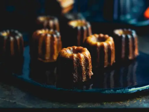 Plusieurs cannelés dorés, parfaits pour la conservation, sont disposés sur une assiette sombre.