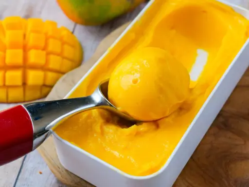 Une cuillère prélève une boule de sorbet mangue crémeux d'une boîte. Des morceaux de mangue fraîche sont visibles à côté.