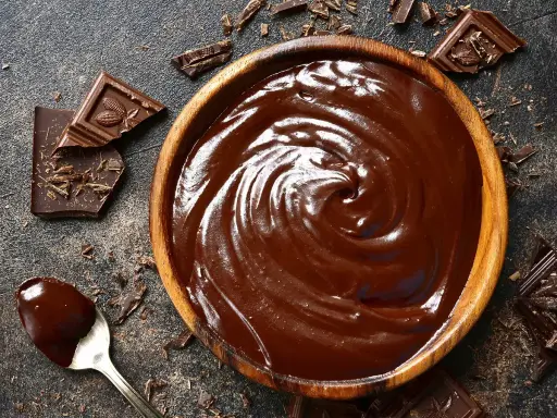 Un bol en bois déborde de chocolat fondu, prêt à faire du chocolat. Des éclats de chocolat noir et une cuillère en argent complètent la scène gourmande.