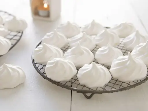 Douces meringues blanches, prêtes pour la cuisson. Elles reposent sur des grilles, évoquant la douceur et la légèreté.