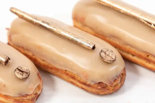 Trois éclairs gourmands, glacés à la crème patissière café, ornés d'une touche dorée et d'un grain de café scintillant.
