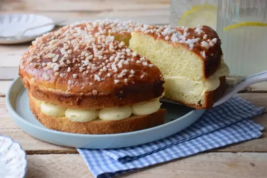 Un délicieux gâteau tropézien, garni de crème onctueuse et saupoudré de sucre perlé. Une part est servie sur une pelle à gâteau.