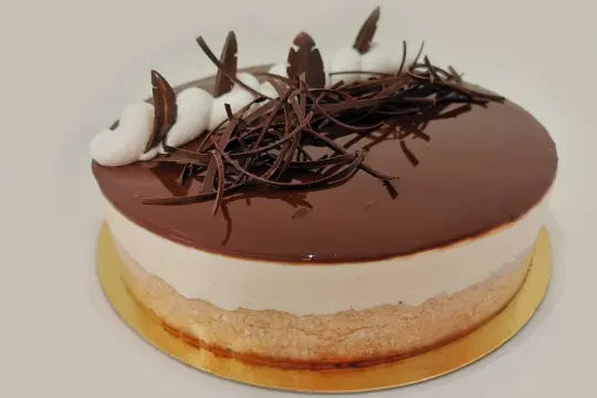Gâteau rond avec glaçage au chocolat brillant, décoré de copeaux de chocolat et de touches de crème. Une délicieuse bavaroise vanille se cache sous le glaçage.