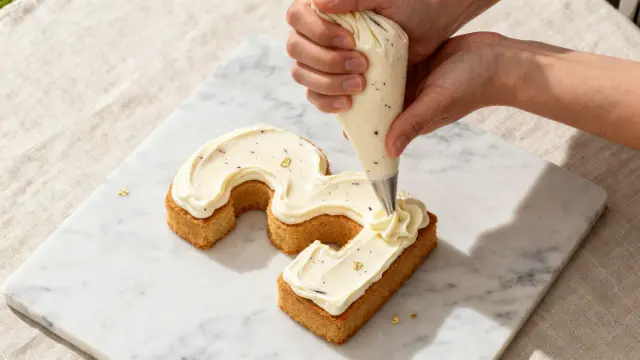 Une main décore un gâteau en forme de chiffre 3 avec de la crème montée et des paillettes dorées.