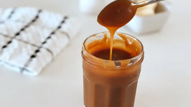 Une cuillère verse un caramel beurre salé liquide dans un pot en verre. Une serviette à carreaux est à gauche.