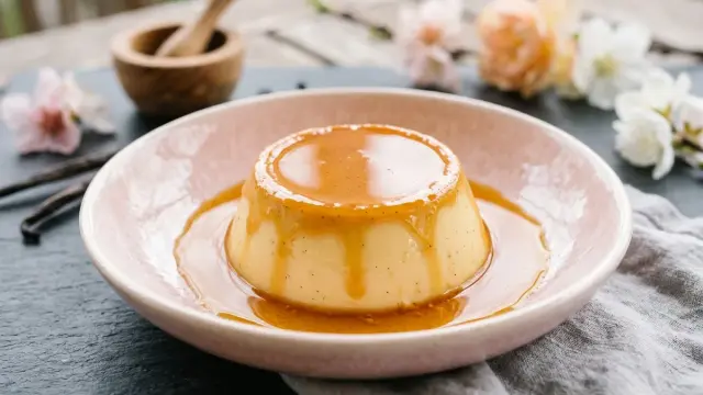 Un délicieux crème caramel, nappé d'une sauce ambrée, repose dans une assiette rose. Des gousses de vanille et des fleurs décorent la scène.