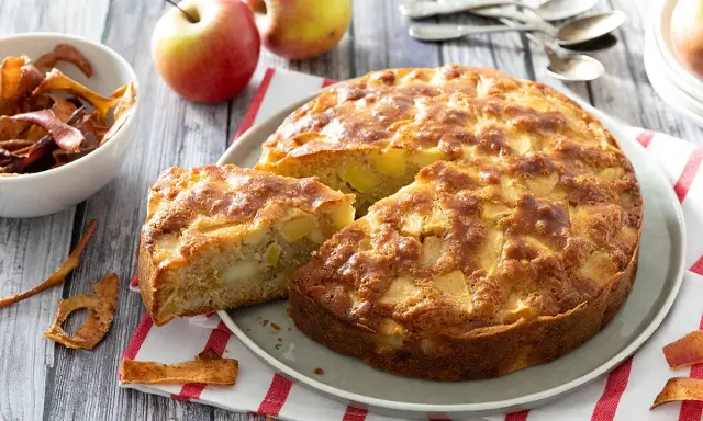 Un gâteau aux pommes super moelleux, doré et appétissant, avec des morceaux de pommes visibles. Des pommes fraîches et des chips de pommes sont disposées autour.