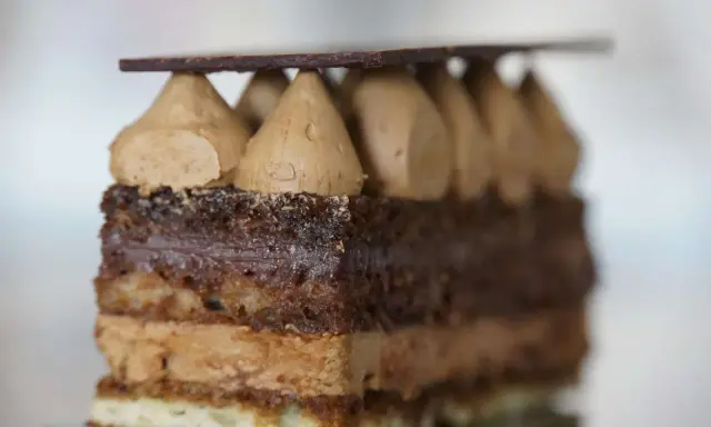 Une tranche de gâteau Opéra, avec sa composition de couches de biscuit Joconde, ganache au chocolat et crème au café, surmontée de dômes de crème et d'une fine plaque de chocolat.