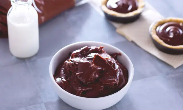 Un bol de ganache chocolat au lait onctueuse, prêt à garnir des tartelettes. Du lait et une poche à douille sont à proximité.
