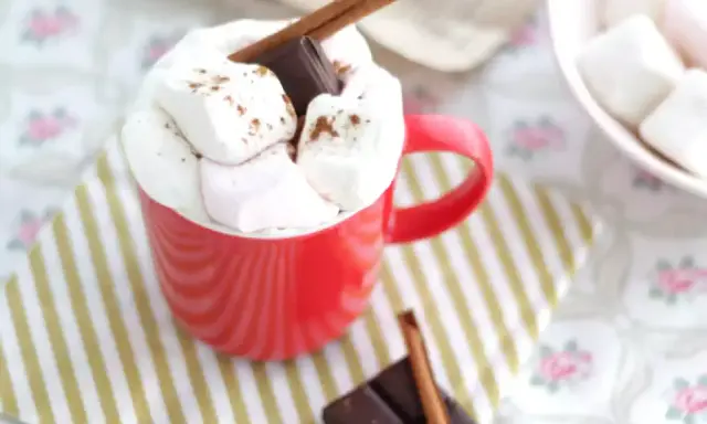 Une tasse rouge débordant de guimauves et de chocolat, parfaite pour une recette guimauve chocolat réconfortante. Un bâton de cannelle et des carrés de chocolat complètent cette gourmandise.