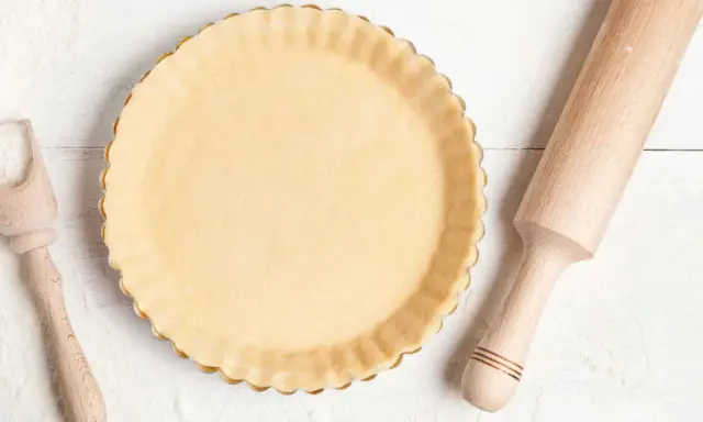 Pâte à tarte crue dans un moule, prête pour le temps de cuisson. Rouleau à pâtisserie et cuillère en bois à proximité.