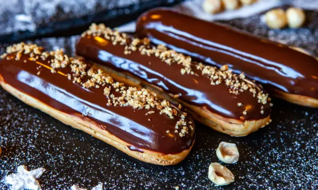 Trois éclairs au chocolat brillants, saupoudrés de noisettes concassées, sur un fond sombre parsemé de sucre glace.
