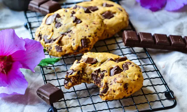 Gros plan sur des cookies chocolat faits maison, un est croqué, révélant un cœur fondant. Des morceaux de chocolat et une fleur d'hibiscus violette complètent la scène.