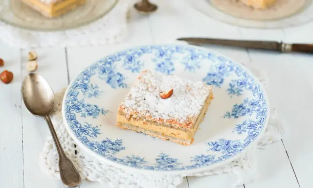 Un carré de gâteau russe, saupoudré de sucre glace et garni d'une noisette, sur une assiette ornée de bleu.