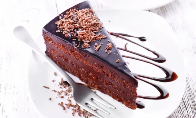 Une part de gâteau aux chocolats, nappée de glaçage brillant et garnie de copeaux de chocolat, est servie sur une assiette blanche avec un filet de sauce chocolat.