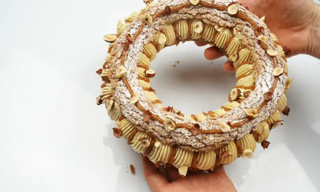 Un Paris-Brest craquelin, orné de noisettes et de caramel, est tenu par des mains.