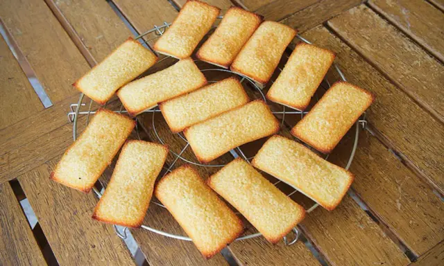 Douze biscuits financiers dorés, disposés sur une grille de refroidissement, sur une table en bois.