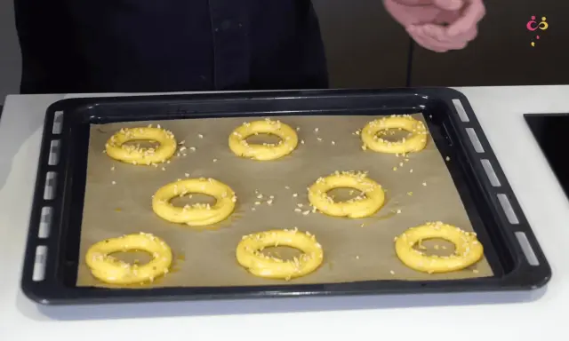 Huit ronds de pâte à choux, prêts à devenir un délicieux Paris-Brest, sont saupoudrés d'amandes effilées sur une plaque de cuisson.