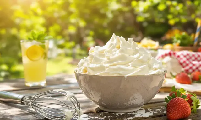 Chantilly aérienne dans un bol, fouet, fraises et limonade sur une table en bois ensoleillée.
