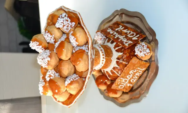 Une pièce montée de choux garnis de crème, nappés de caramel et décorés de perles blanches. Sur un plateau, d'autres choux et des tuiles aux amandes portent des inscriptions.