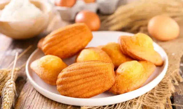 Des madeleines dorées, parfaites pour une recette gâteau madeleine meilleur pâtissier. Des œufs et de la farine en arrière-plan.