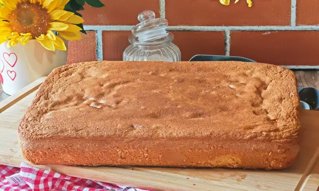Une génoise rectangulaire dorée, parfaite pour 15 personnes, attend d'être garnie. Des fleurs jaunes et un fond de briques rouges créent une ambiance chaleureuse.