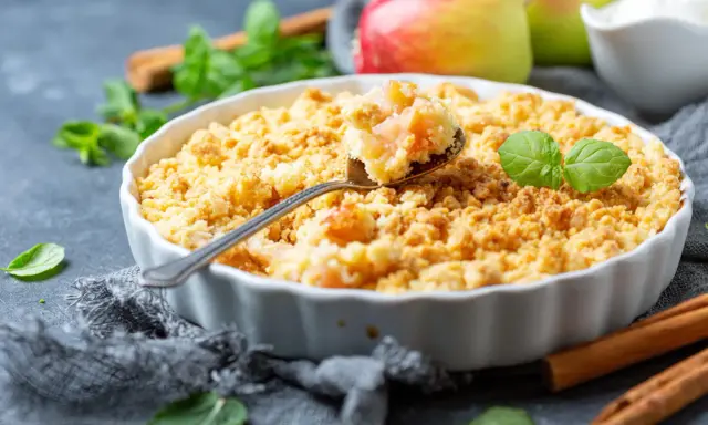 Un délicieux crumble aux pommes à l'ancienne, garni de feuilles de menthe fraîche, avec des pommes et des bâtons de cannelle en arrière-plan.