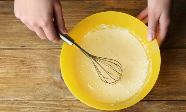 Mains préparant la pâte à crêpes dans un bol jaune. La pâte est prête pour son temps de repos.
