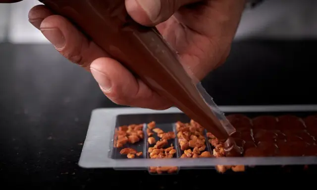 Main d'un artisan versant du chocolat fondu dans un moule pour créer une tablette de chocolat maison garnie de noisettes caramélisées.
