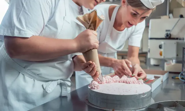 Deux pâtissières décorent un gâteau rose lors d'un cours de pâtisserie en ligne. L'une utilise une poche à douille, l'autre des fruits.