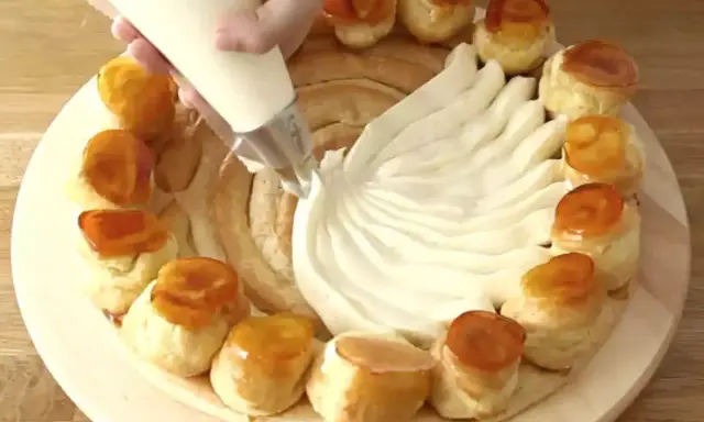 Une main garnit une couronne de choux avec de la crème chiboust, créant un dessert spectaculaire.