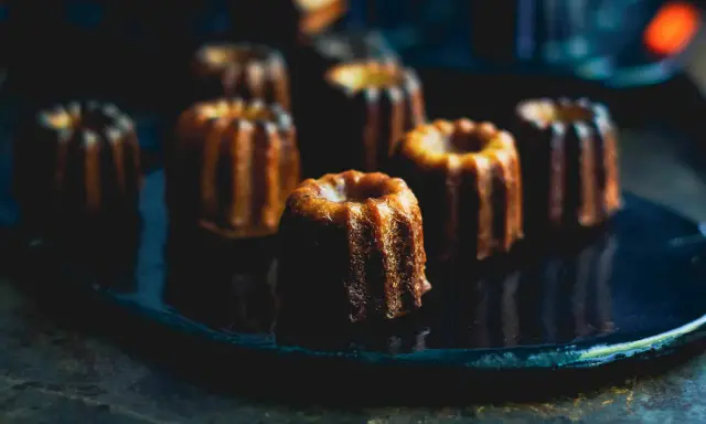Plusieurs cannelés dorés, parfaits pour la conservation, sont disposés sur une assiette sombre.