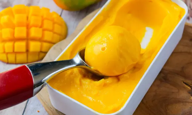 Une cuillère prélève une boule de sorbet mangue crémeux d'une boîte. Des morceaux de mangue fraîche sont visibles à côté.