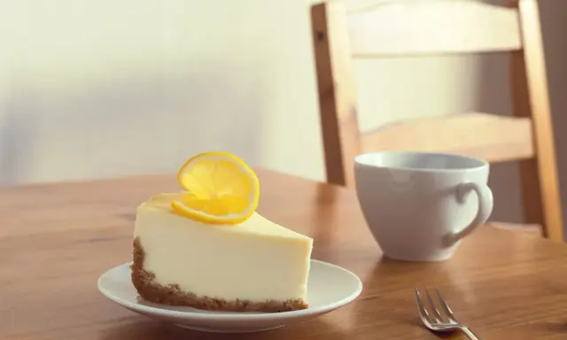 Une part de le meilleur cheesecake au citron, garnie d'une tranche de citron, sur une assiette blanche, à côté d'une tasse et d'une fourchette sur une table en bois.
